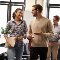 Two young diverse business people male and female coworkers talking discussing project and smiling while walking down the modern office